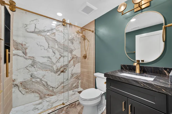 Bathroom featuring a frameless glass shower enclosure with gold-tone hardware, large format veined wall tiles, a dark vanity with integrated sink, a dark countertop, and a matching gold-tone wall-mounted faucet