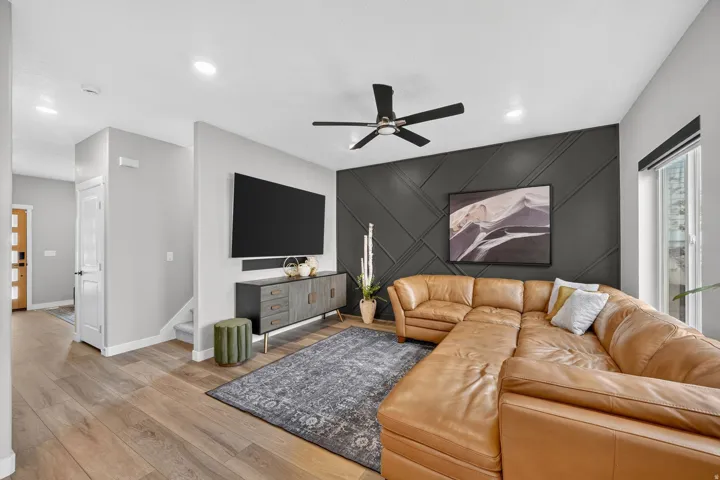 Spacious living area featuring light wood-finish flooring, a dark accent wall with geometric paneling, and a ceiling fan with integrated lighting