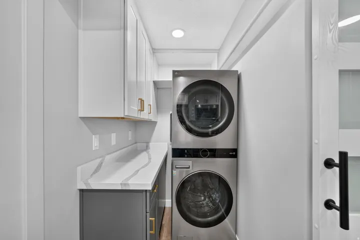 Dedicated laundry area featuring stacked stainless steel appliances, a white and gray veined countertop, and white upper cabinetry with gold-finish hardware