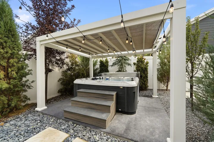 Outdoor spa area featuring a dark-toned hot tub with integrated steps, shaded by a white pergola with string lighting