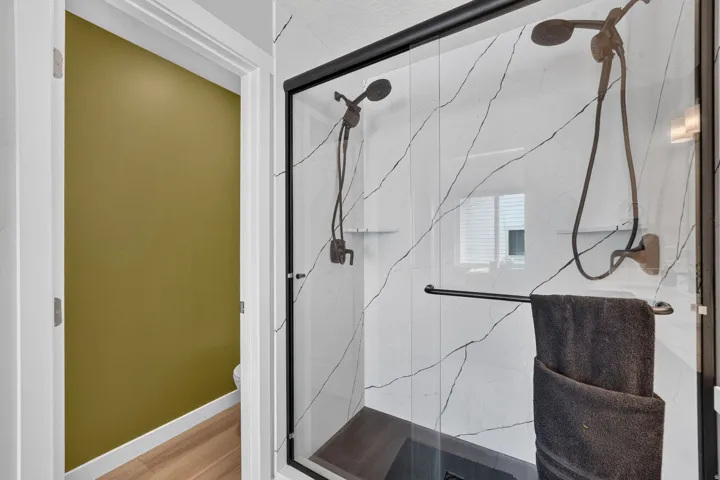 Spacious walk-in shower featuring clear glass enclosure with matte black framing, white tile with dark veining, two bronze-finish showerheads, and a dark tile shower pan