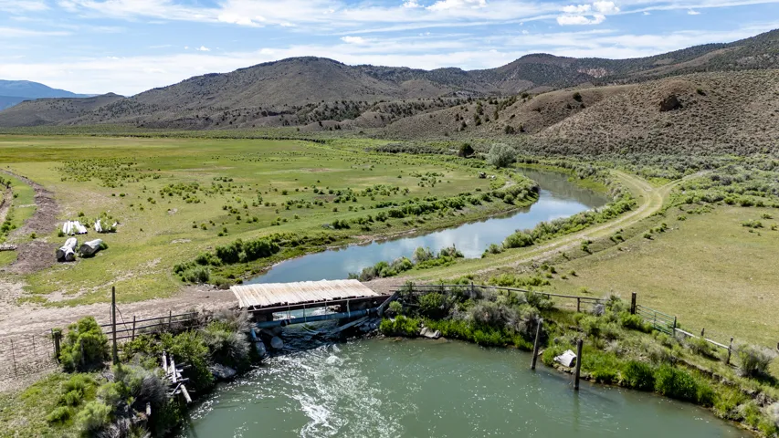 Expansive natural landscape featuring a river, verdant fields, rolling hills, and a wooden bridge
