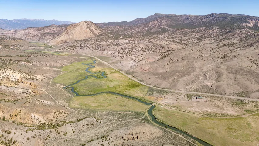Expansive rural landscape featuring a winding river, verdant riverbanks, surrounding arid hills, distant mountain ranges, and an unpaved road