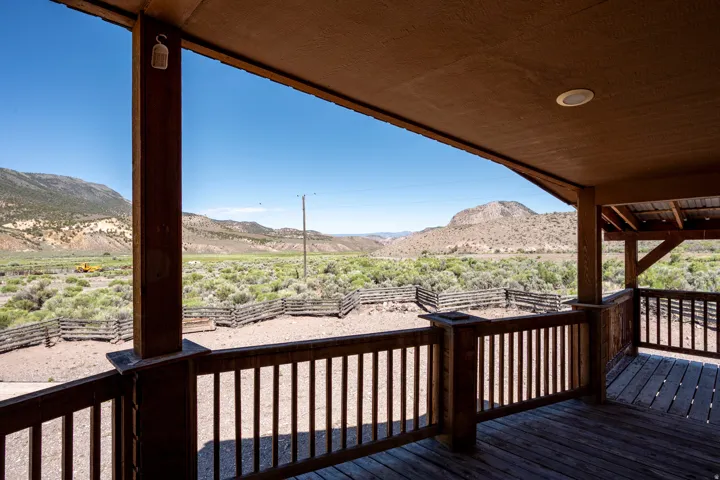 Expansive covered deck with wood flooring and railing, offering views of the surrounding mountainous landscape, fenced corrals, and natural vegetation