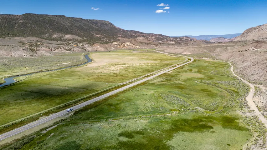 Expansive green valley featuring a winding river, a paved highway, rugged mountain backdrops, and a clear blue sky