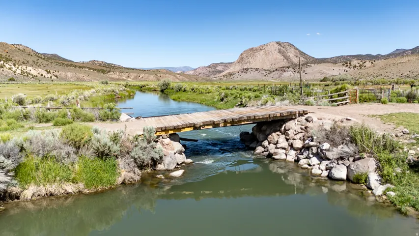 Rustic wooden bridge crossing a flowing river, surrounded by expansive green fields and distant mountains
