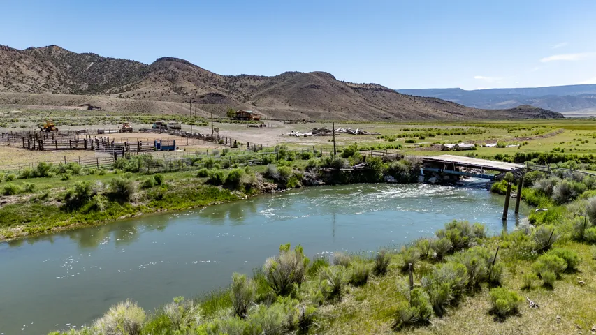 Expansive property featuring a river, mountainous backdrop, a wooden bridge, and livestock pens