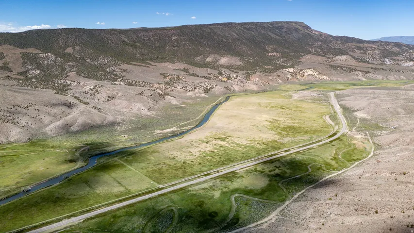 Expansive mountainous landscape featuring a river, verdant fields, a paved road, and a dirt path