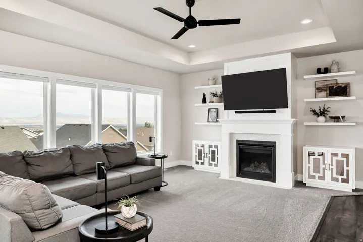Living area featuring a tray ceiling with recessed lighting and a ceiling fan, a prominent fireplace with a mantel, built-in shelving, and large windows with surrounding landscape