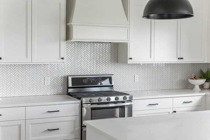 Kitchen featuring white shaker cabinetry, herringbone tile backsplash, stainless steel range, light-toned countertops, and a built-in range hood