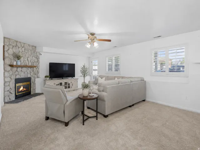 Living area featuring a stone-clad fireplace with a wood mantel, light-toned carpeting, white walls, and multiple windows with plantation shutters