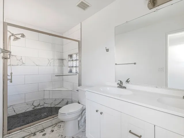 Bathroom featuring a dual sink vanity with white cabinetry, a frameless mirror, and a glass-enclosed shower with tile surround, a built-in bench, and a glass block window