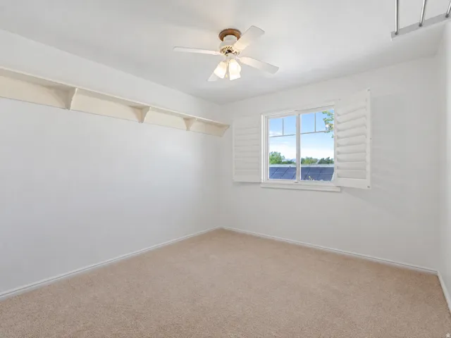 Carpeted interior room featuring a window with white louvered shutters, built-in wall shelving, a ceiling fan with integrated lighting, and white baseboards