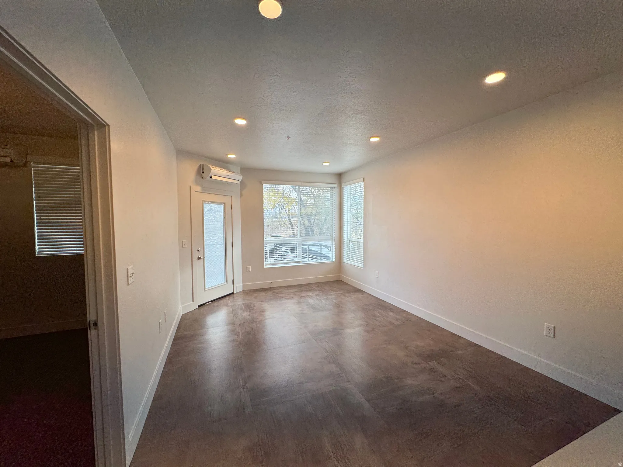 Spacious room featuring wood-finish flooring, recessed lighting, multiple windows, a glass-paneled door, and a wall-mounted mini-split system