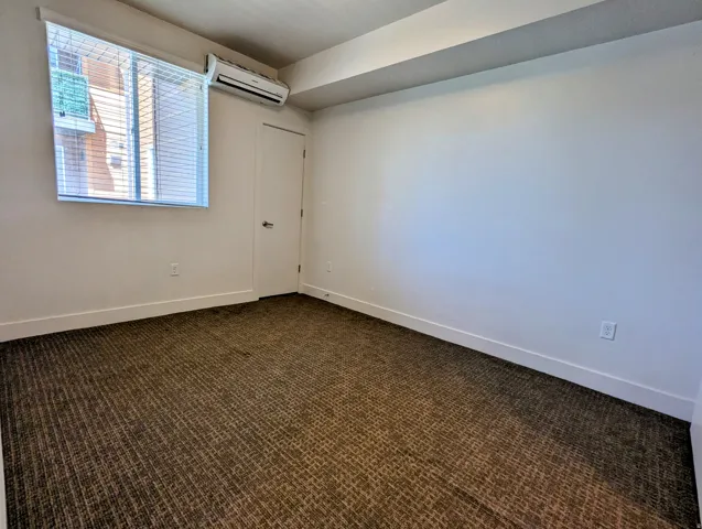 White walls with white baseboards and trim, brown patterned carpeting, a single window with horizontal blinds, and a wall-mounted mini-split HVAC unit