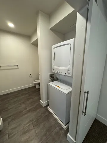 Compact laundry area featuring a stacked washer and dryer unit, dark tone tile flooring, a white barn door, recessed lighting, and a chrome towel bar