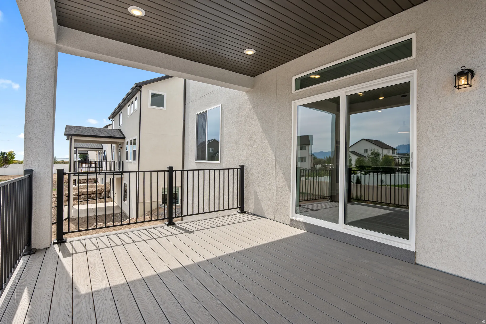Spacious covered deck featuring composite decking, black metal railings, recessed lighting, and a large sliding glass door with a transom window