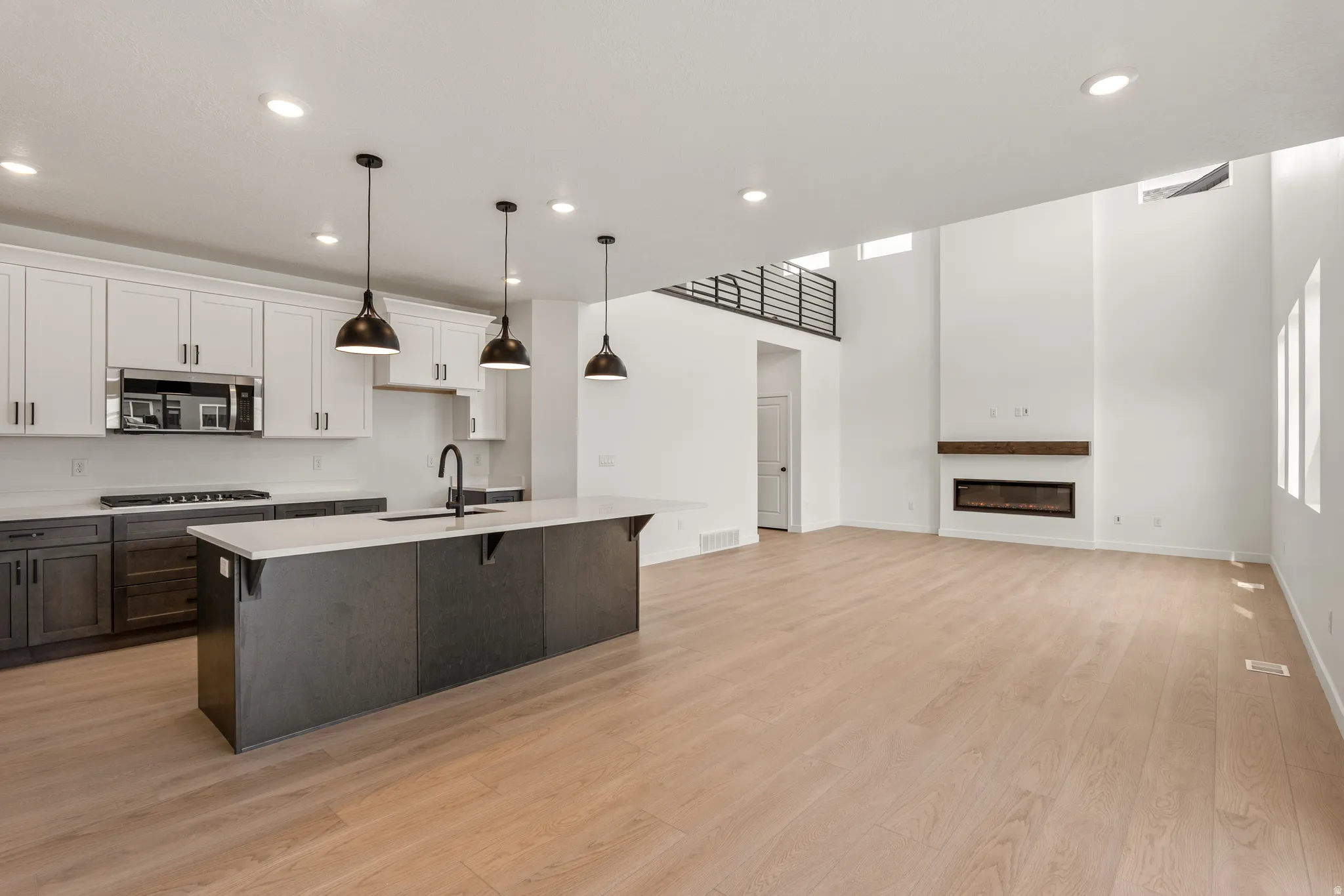 Open-concept main level featuring light wood-finish flooring, a contemporary linear fireplace with a wood mantle, and an upper-level metal railing overlooking the space