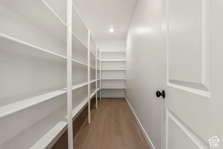 Walk-in pantry featuring extensive white shelving, recessed ceiling lighting, a paneled door with a black knob, and wood-finish flooring
