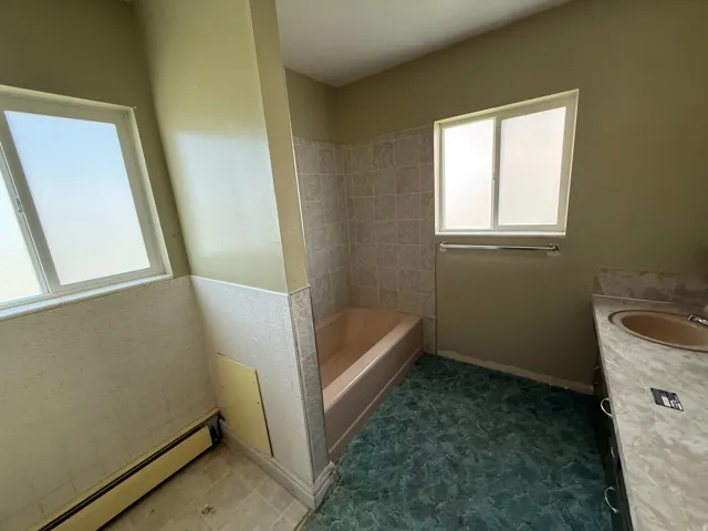 Bathroom featuring two windows with frosted panes, a built-in bathtub with tiled surround, a vanity with an integrated sink and countertop, and a baseboard heater