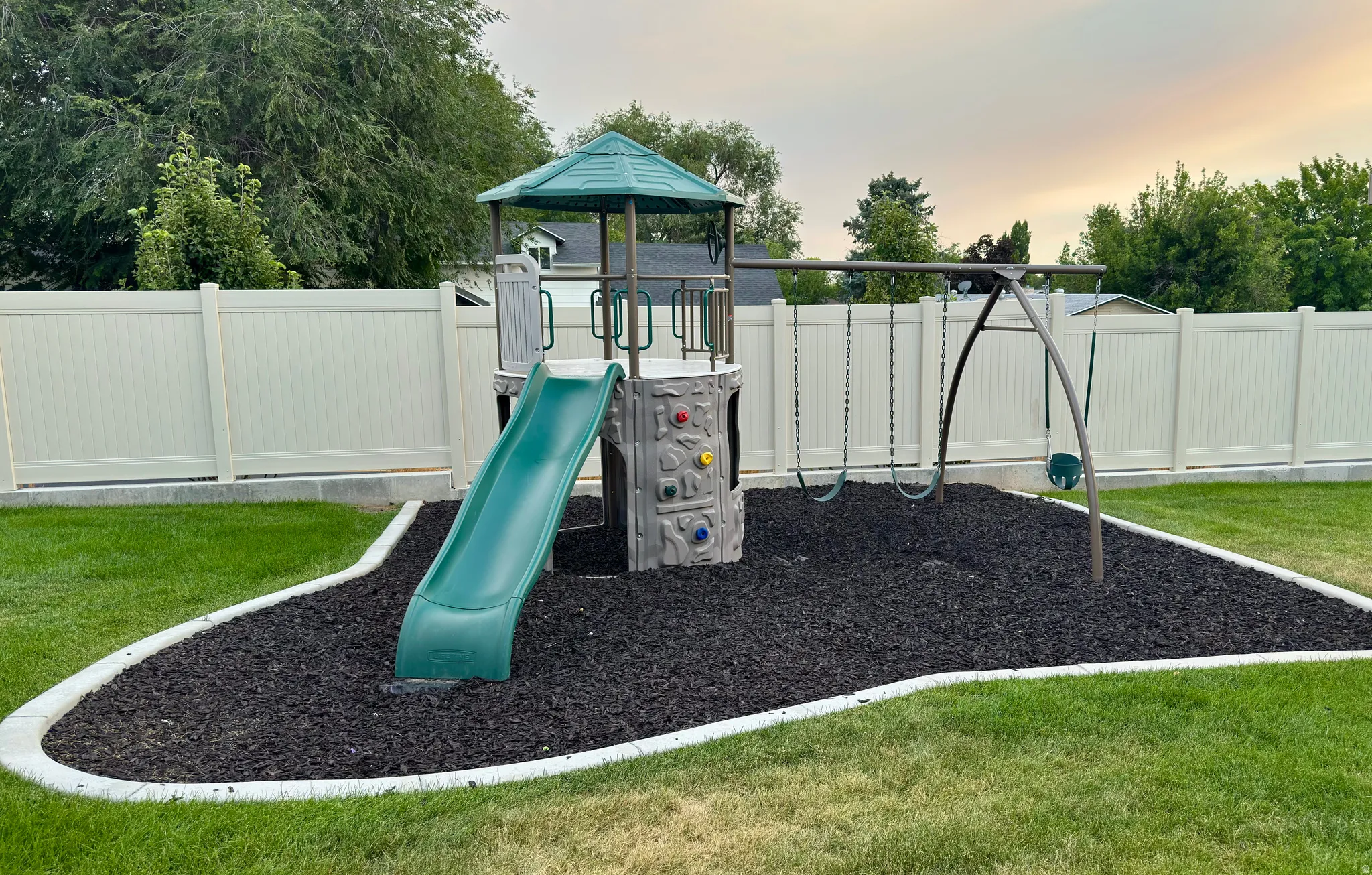 Common playground area featuring a multi-level playset with slide and rock climbing wall, a swing set, and a perimeter of black rubber mulch