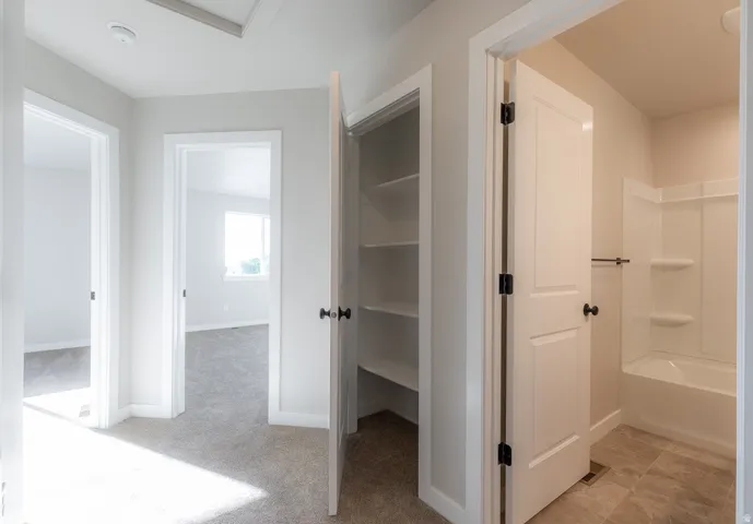 Upstairs hallway featuring light neutral walls and plush carpeting, with a white door opening to a bathroom with a white tub-shower combination and tiled flooring