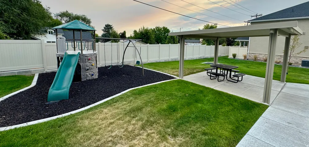 Common playground and covered picnic areas featuring a covered patio, a separate play structure area with rubber mulch, and a manicured lawn enclosed by a white privacy fence