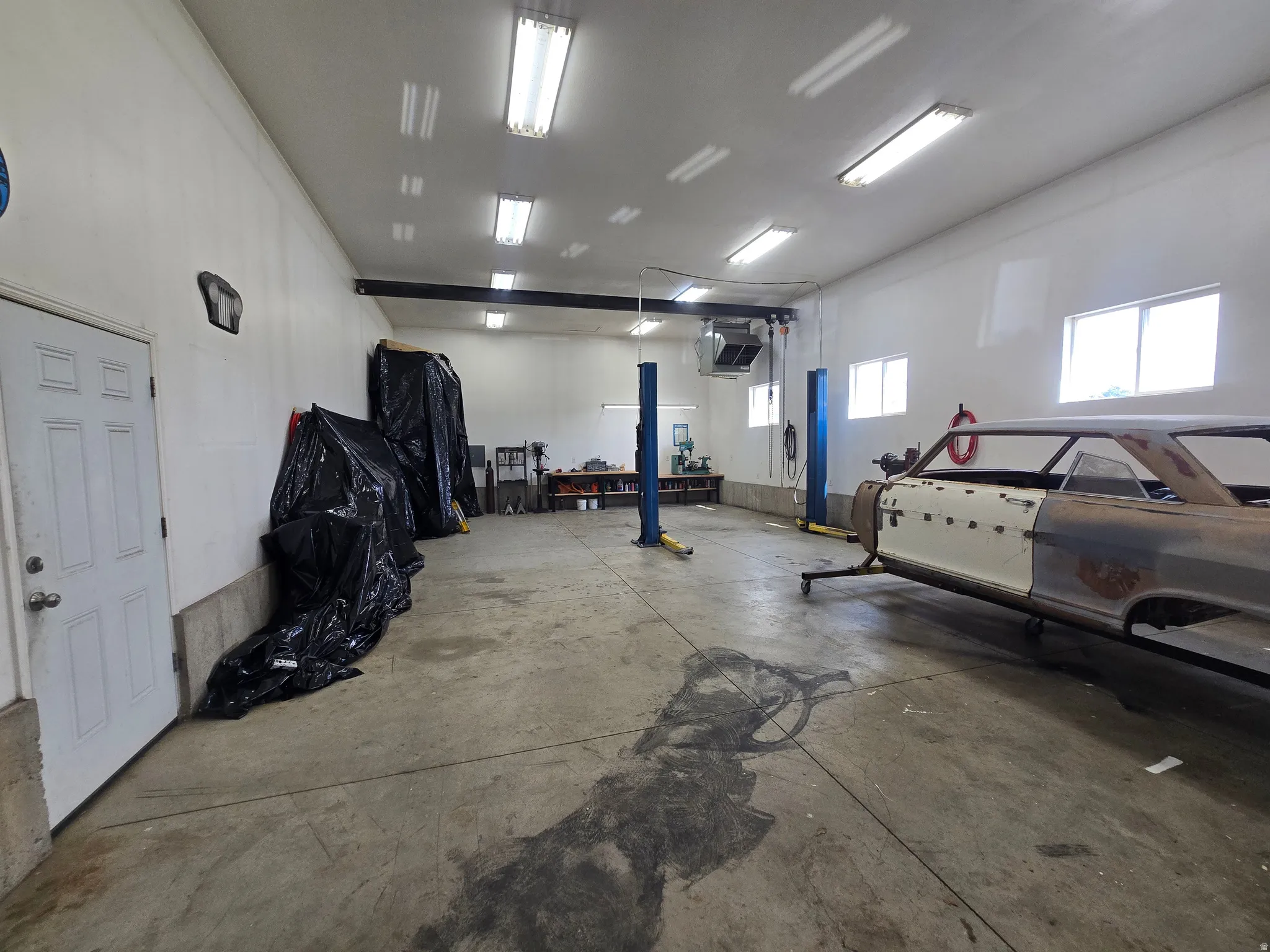 Spacious garage workshop featuring concrete flooring, multiple windows, fluorescent lighting, and a vehicle lift