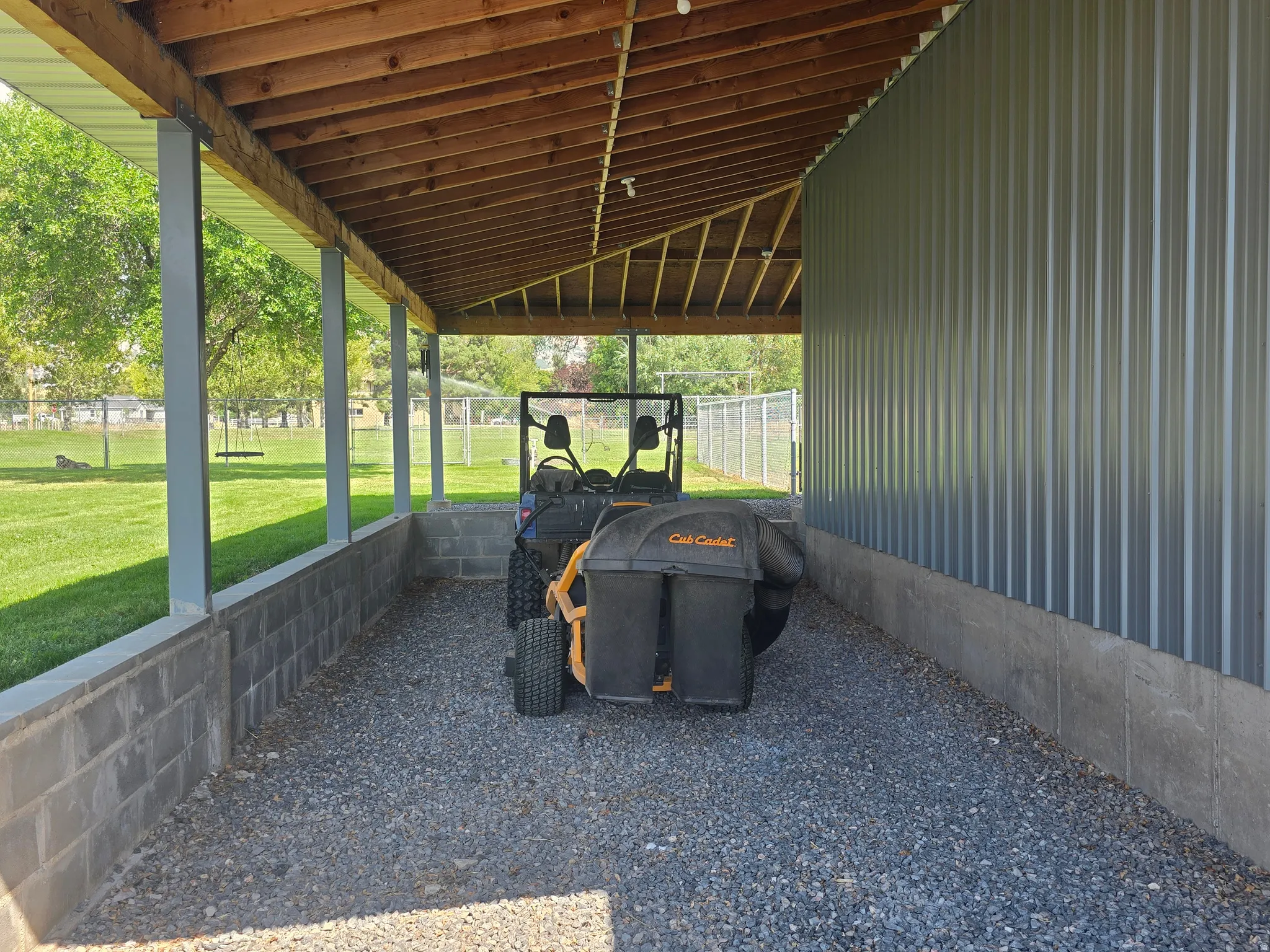 Covered outdoor storage area featuring a gravel surface, cinder block perimeter, exposed wood beam ceiling, and corrugated metal siding