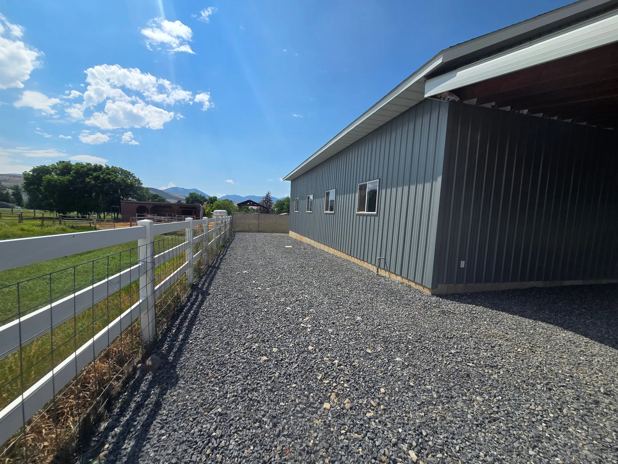 Gravel driveway alongside a metal-clad structure with multiple windows, featuring a white fence bordering a grassy area