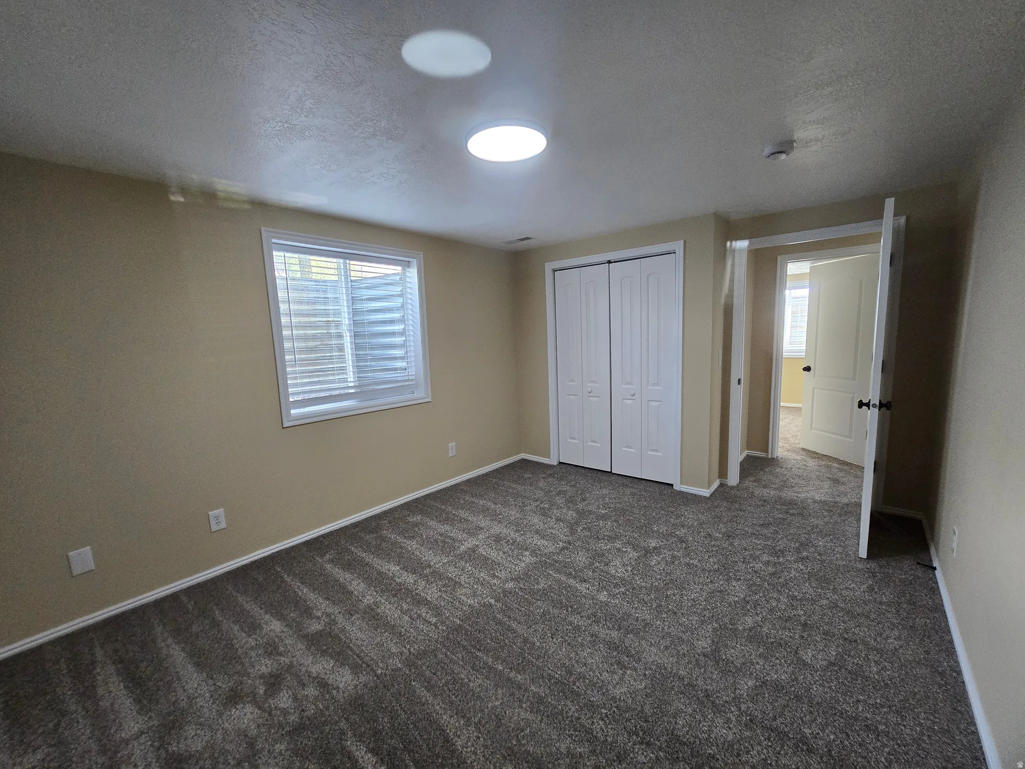 Spacious room featuring neutral wall tones, plush gray carpet, a white window with horizontal blinds, bi-fold closet doors, and recessed ceiling lights