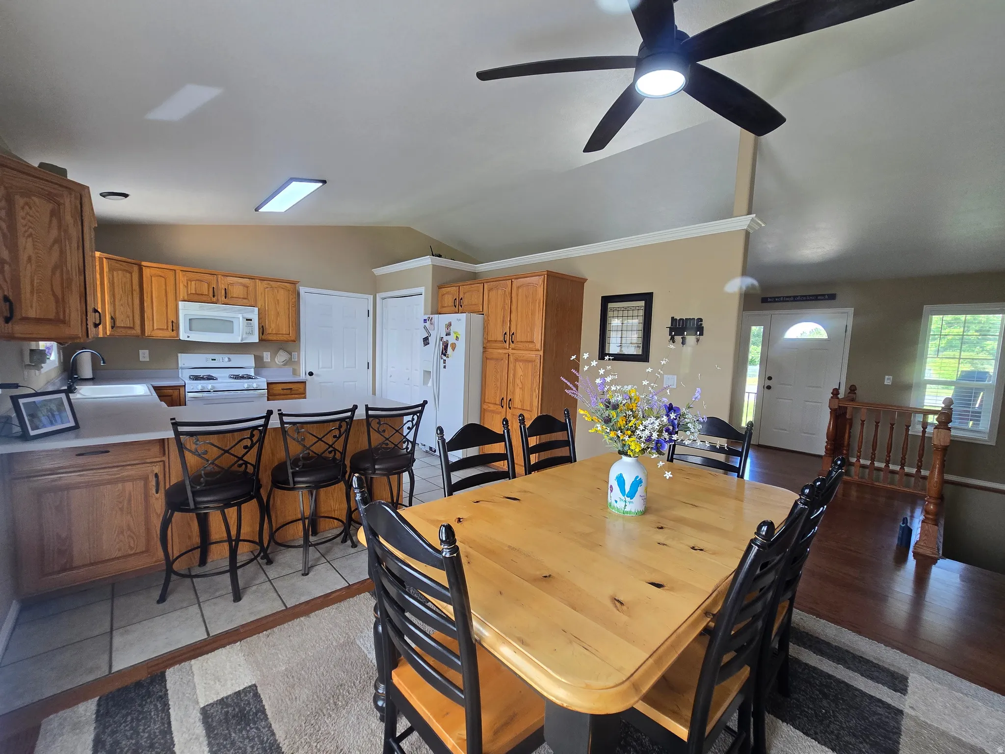 Open-concept kitchen and dining area featuring warm wood cabinetry, a breakfast bar with seating, white appliances, and a vaulted ceiling with a ceiling fan