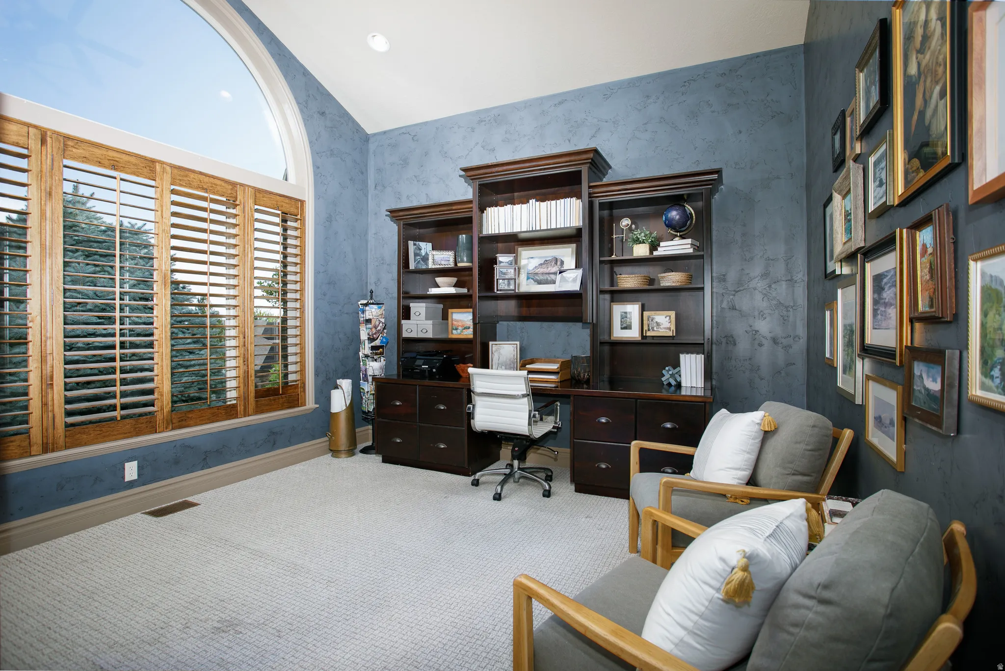 Office space featuring a large arched window with custom wood shutters, built-in dark wood cabinetry, textured blue walls, and light-colored carpet flooring