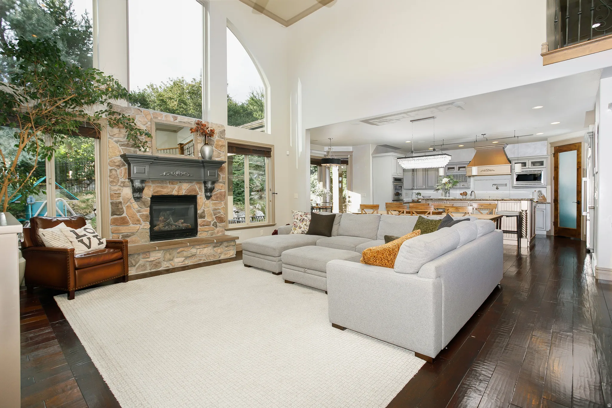 Spacious living area featuring a stone fireplace with a gray mantle, dark wood-finish flooring, and high ceilings with large windows