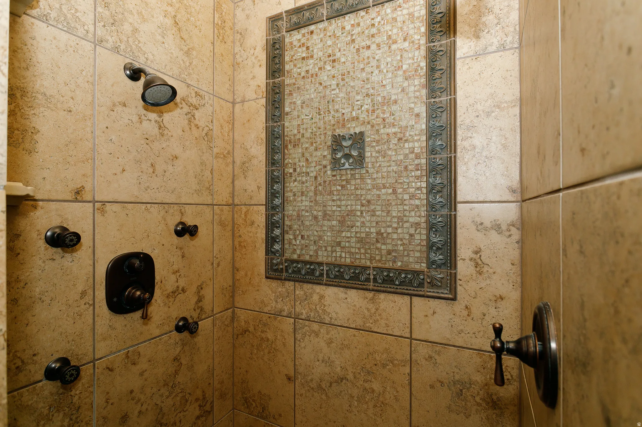 Tiled shower enclosure featuring a mosaic accent panel with decorative trim, bronze-finish fixtures, and a rain shower head