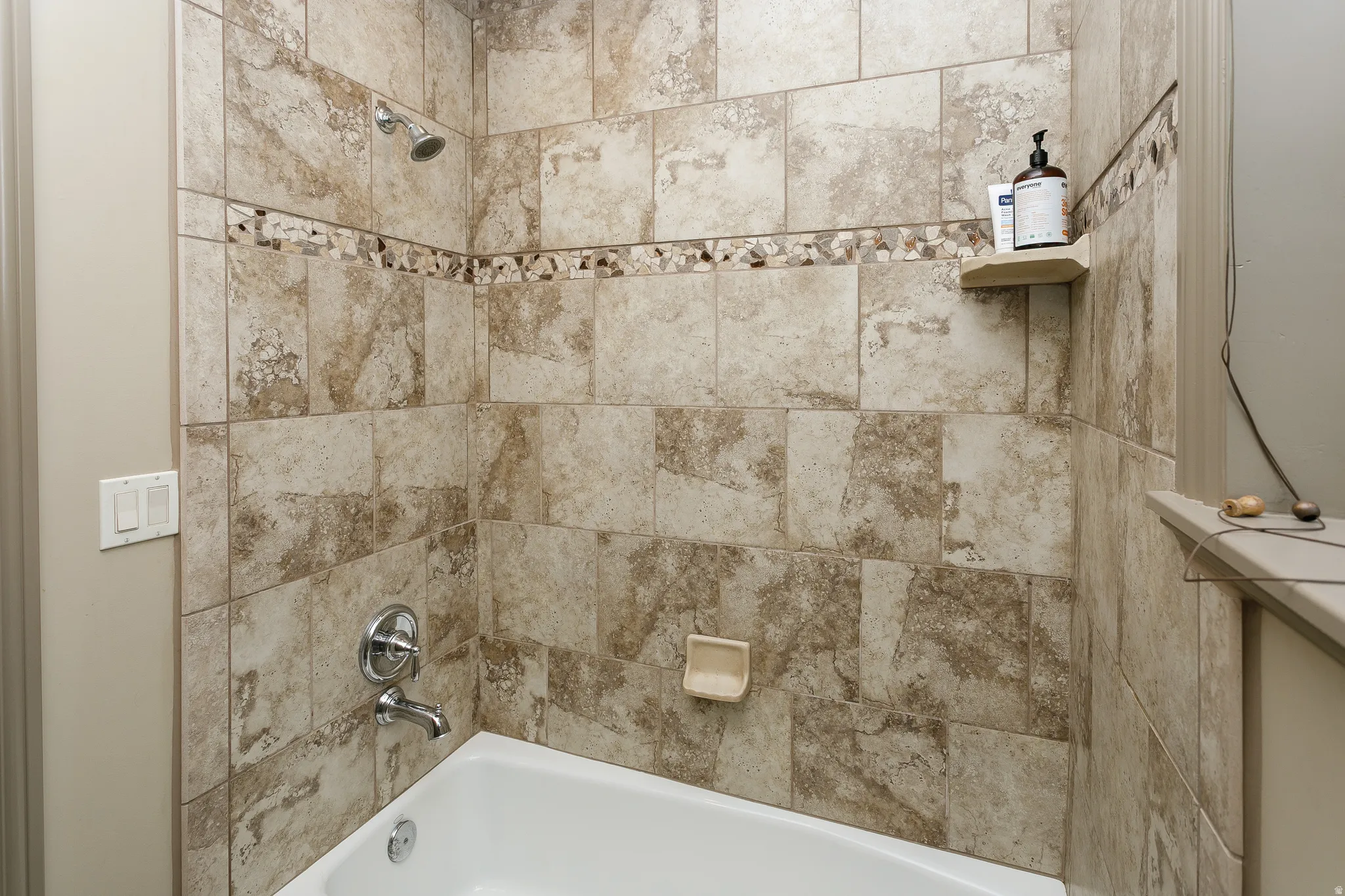 Bathtub and shower combination featuring neutral-toned tile surround with decorative mosaic accent band
