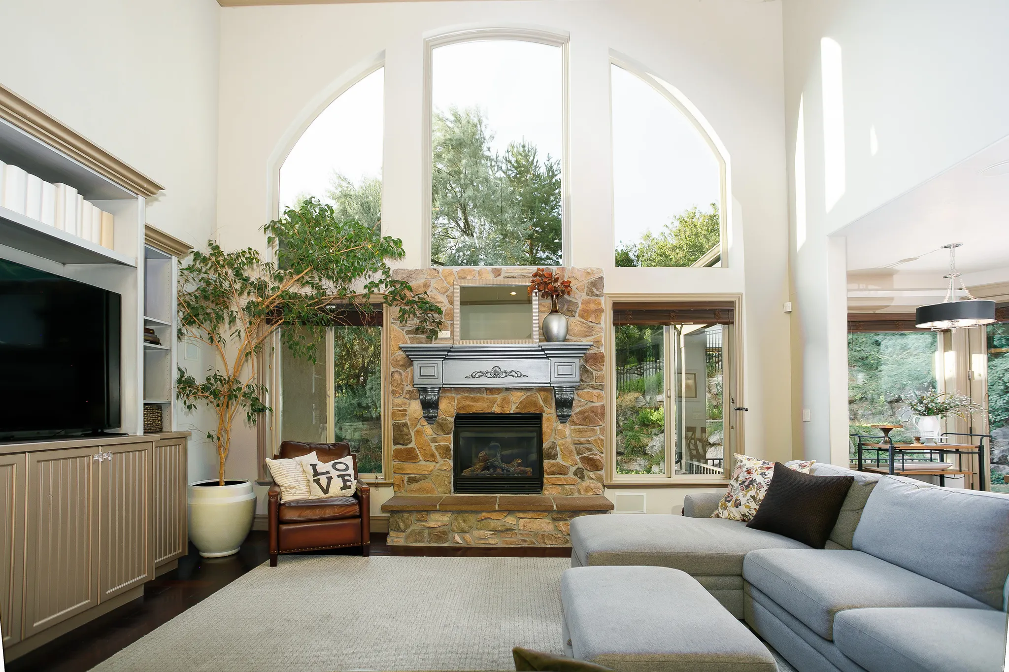 Vaulted living space featuring a stone fireplace with ornate mantel, expansive arched windows, dark wood-finish flooring, and built-in media cabinetry with shelving