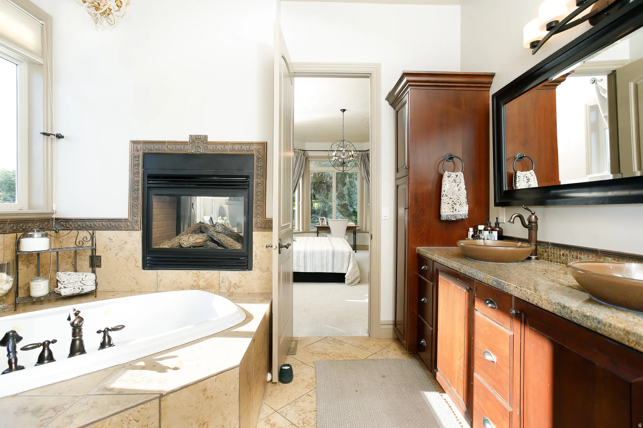 Freestanding soaking tub with a tiled surround, gas fireplace with decorative metal trim, dual vanity with vessel sinks, granite countertop, and custom wood cabinetry