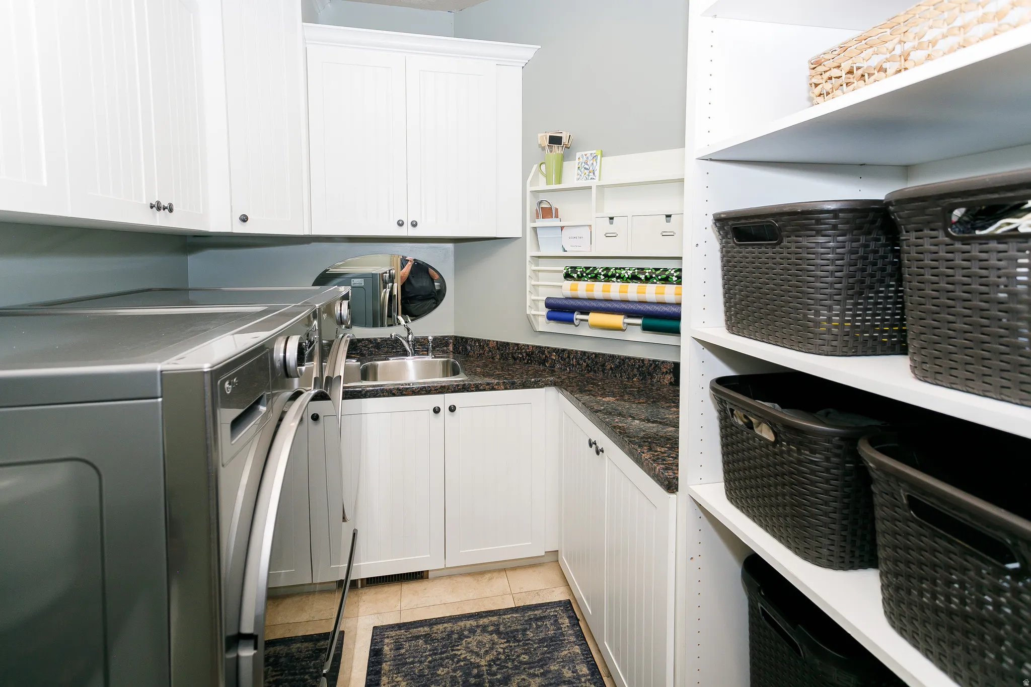 Dedicated utility room featuring white cabinetry, a stainless steel sink, a dark speckled countertop, built-in shelving, and light tile flooring