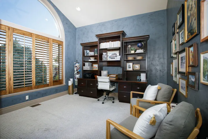 Office space featuring a large arched window with custom wood shutters, built-in dark wood cabinetry, textured blue walls, and light-colored carpet flooring