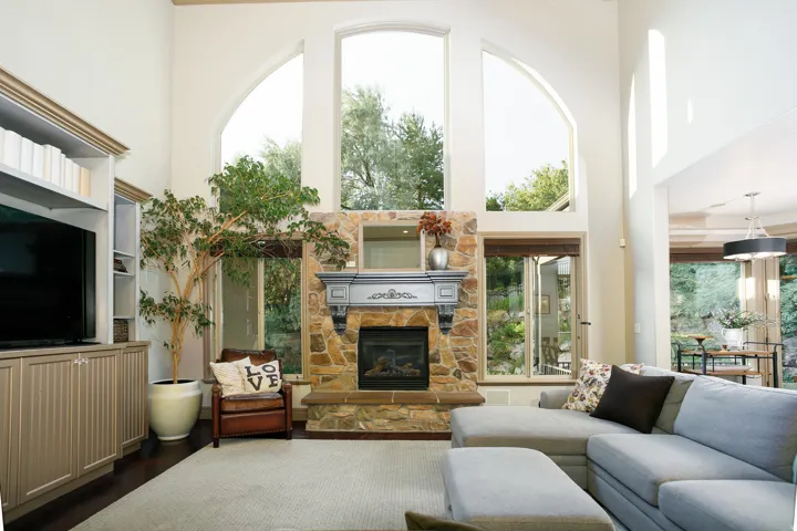 Vaulted living space featuring a stone fireplace with ornate mantel, expansive arched windows, dark wood-finish flooring, and built-in media cabinetry with shelving