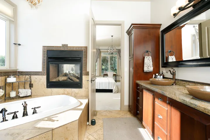 Freestanding soaking tub with a tiled surround, gas fireplace with decorative metal trim, dual vanity with vessel sinks, granite countertop, and custom wood cabinetry