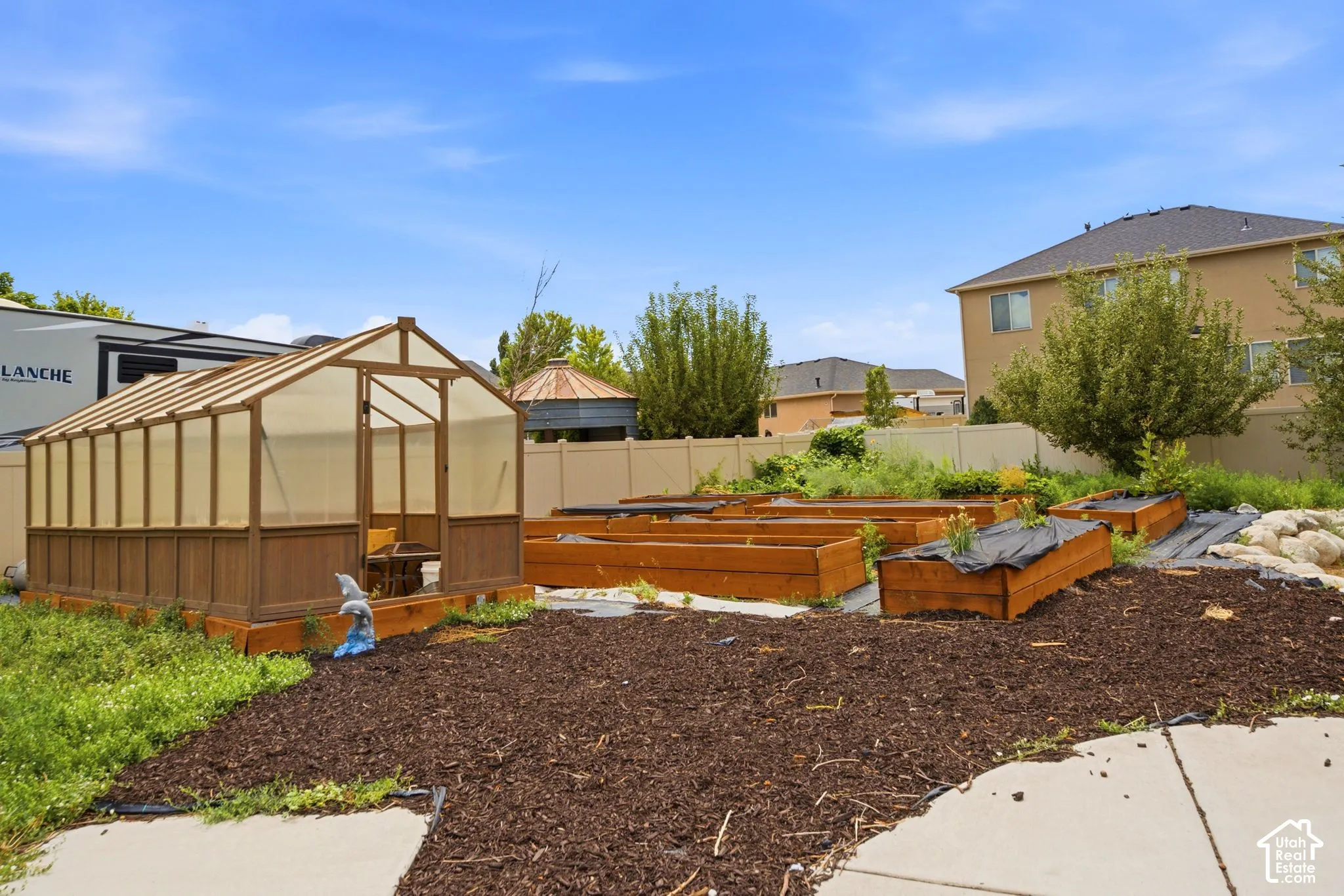 Backyard featuring a greenhouse structure with a wood-finish frame and translucent panels, multiple raised garden beds, and a perimeter fence