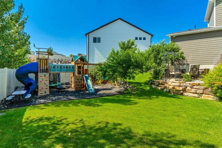 Expansive backyard featuring a lush lawn, a multi-level play structure with slides and climbing walls, a white privacy fence, a natural stone retaining wall, and mature landscaping