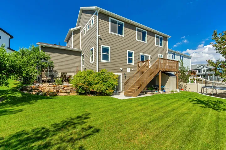 Expansive rear lawn featuring a wooden deck with stairs, a stone retaining wall, and a concrete patio