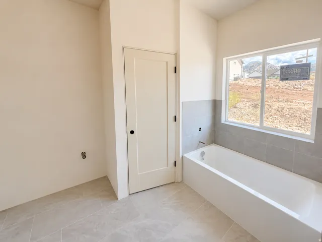 White alcove bathtub with light gray tile surround, set below a large window