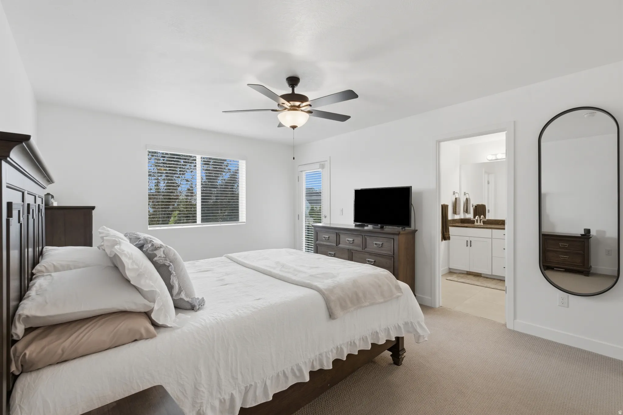 Carpeted bedroom featuring a ceiling fan with light fixture, window with blinds, and an open doorway revealing a bathroom with a white vanity and brown countertop