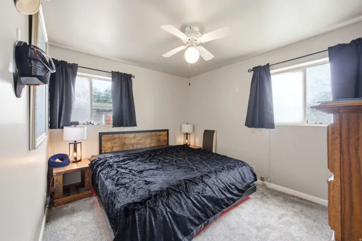 Carpeted room featuring a ceiling fan with integrated lighting, a distressed wood-finish headboard, and two windows with dark drapery