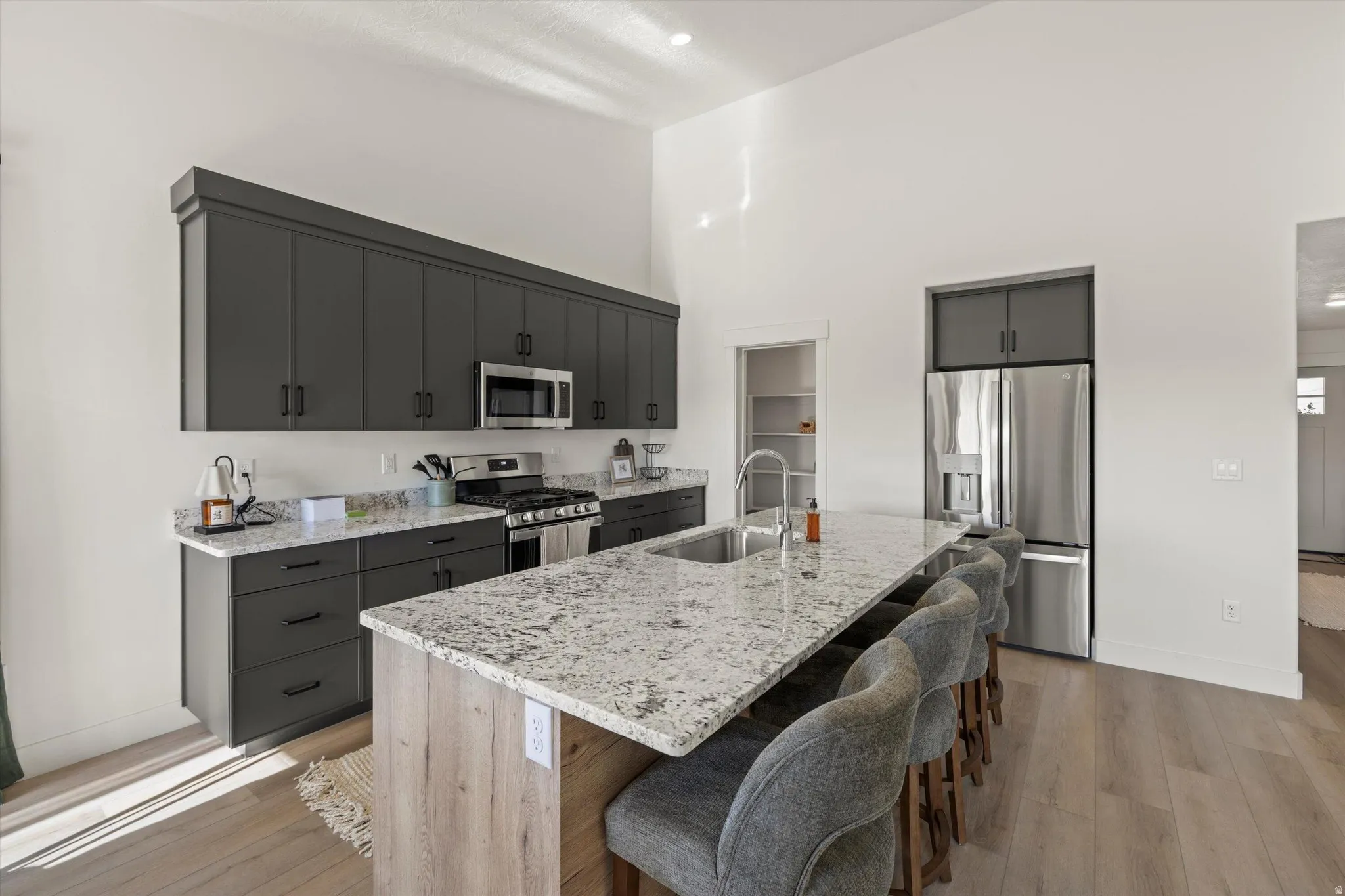Bright kitchen featuring dark gray cabinetry, stainless steel appliances, light-toned wood-finish flooring, a large island with an integrated sink, and light granite countertops