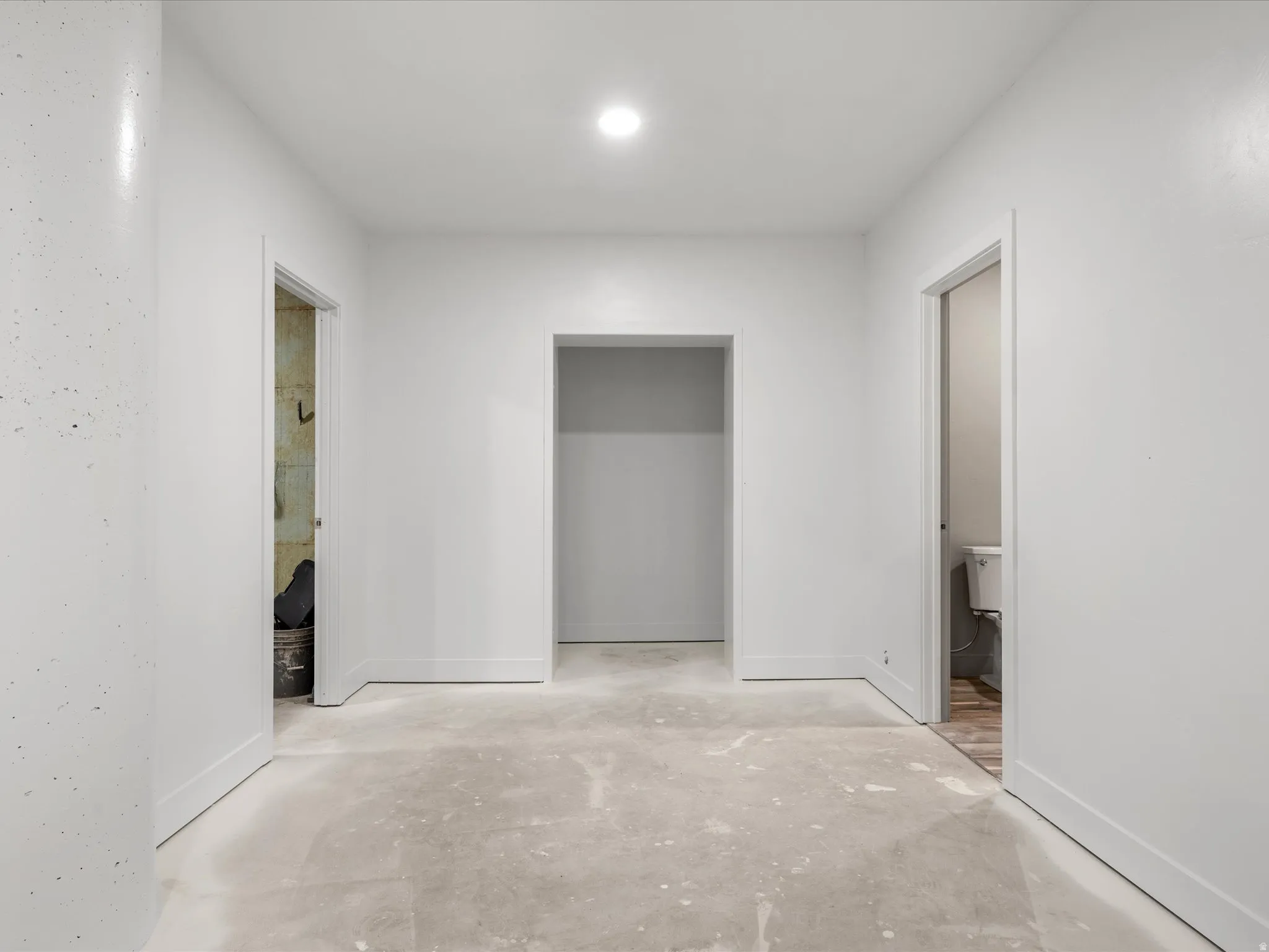Open hall with white walls and baseboards, recessed ceiling lighting, two doorways with white trim, and a concrete subfloor